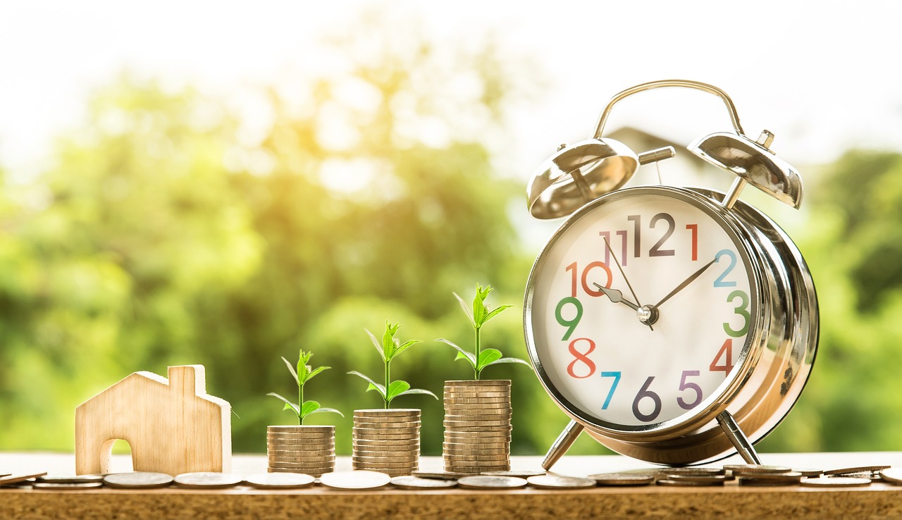 A blurred background image of the sun and green trees with the foreground showing a table with coins in three incrementally taller piles each pile with a small green plant shoot growing out of them. To the left of the coins a small carved piece of wood of a home and on the right of the coins an old traditional alarm clock with two bells on top either side.
